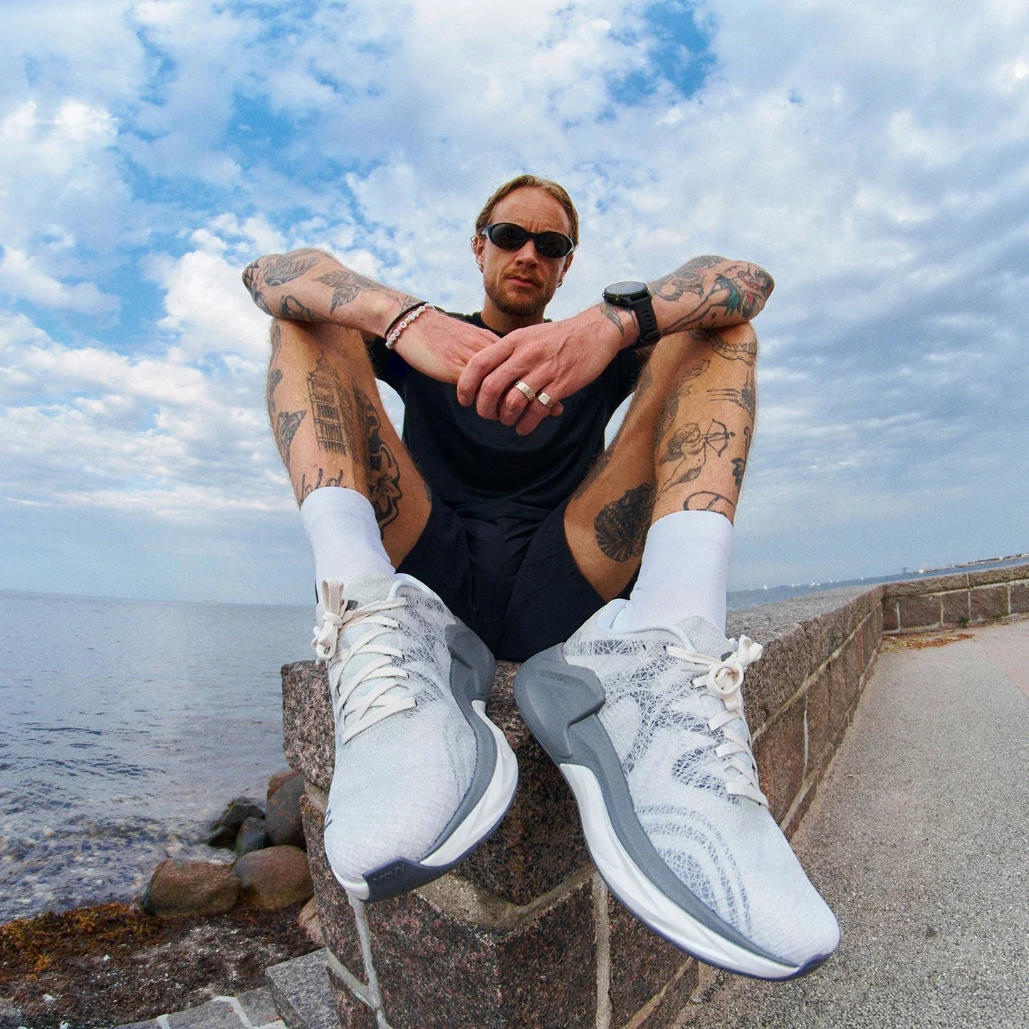 A tattooed man with sunglasses sits on a stone seawall by the ocean, wearing a black tank top and white MOVV running shoes. His arms rest on his knees in a relaxed pose, with extensive tattoo work visible on both arms and legs. The sky behind him is partly cloudy and blue, with water and coastline visible in the background.