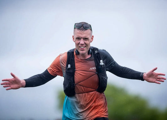 **Alt text:** Smiling male ultrarunner wearing a MOVV shirt and hydration vest with arms outstretched, finishing a race in rainy weather.